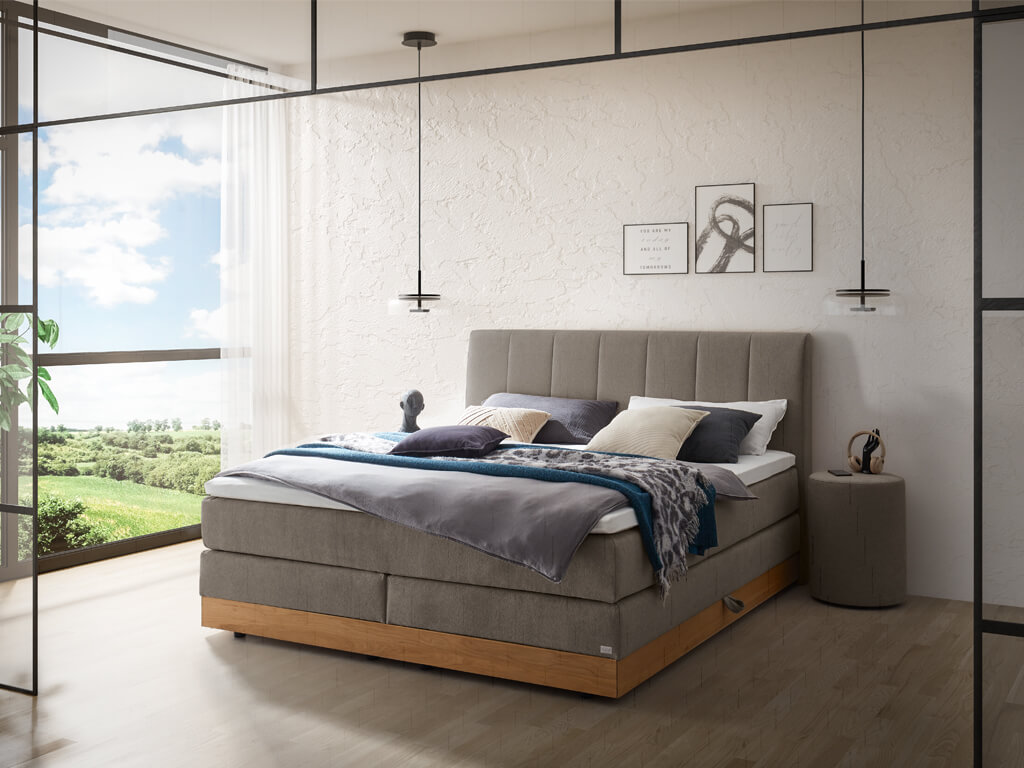 Ein modern eingerichtetes Schlafzimmer mit einem grauen Boxspringbett mit Holzdetails und einem vertikal gesteppten Kopfteil.