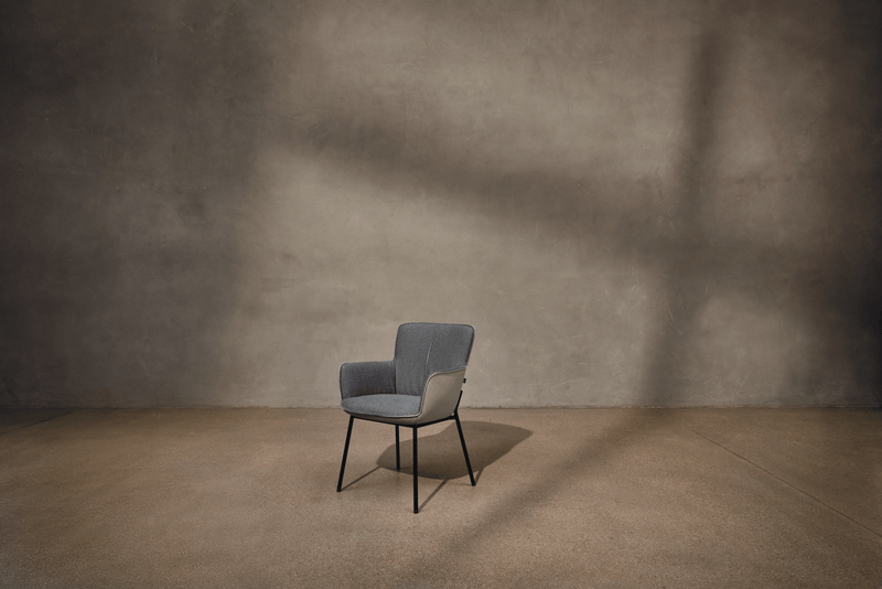 Ein eleganter grauer Stuhl mit schwarzen Beinen, platziert vor einer neutralen Wand mit Licht- und Schatteneffekten.