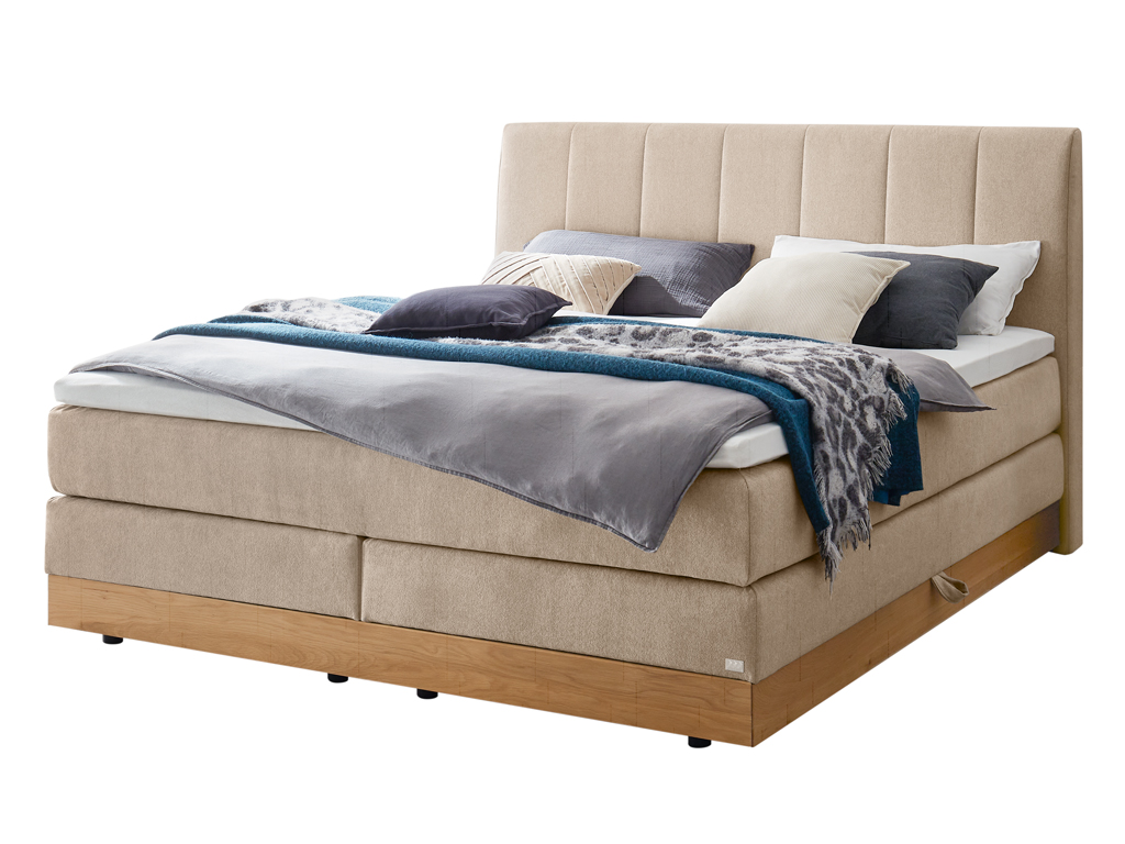 Ein Boxspringbett in beigem Stoff mit einem vertikal gesteppten Kopfteil und einem Holzbettrahmen, freigestellt auf weißem Hintergrund.