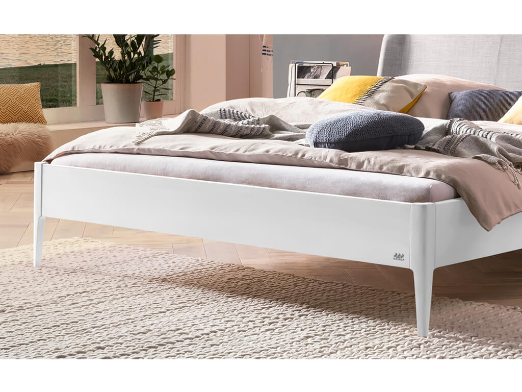 Eine Nahaufnahme des unteren Teils eines Bettes Pienza in Weiß-deckend-lackiert. Das Bett hat schlanke, konische Füße und steht auf einem hellen, gewebten Teppich. Im Hintergrund sind ein großes Fenster, Zimmerpflanzen und ein Teppich zu sehen.