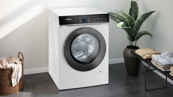 Eine geschlossene Siemens iQ700 Waschmaschine im Raum, platziert neben einem großen Blumentopf und einem Beistelltisch mit Wäsche.