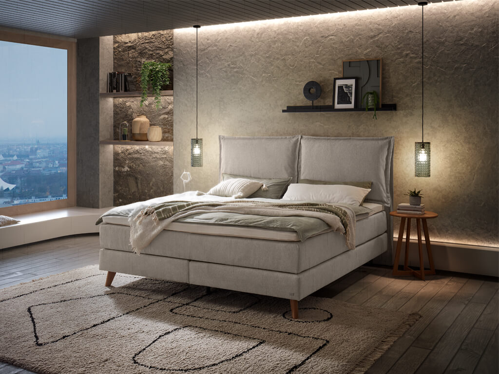 Ein Hasena Boxspringbett Pronto Ricarda Roy mit einem hellgrauen Stoffbezug, das sich harmonisch in ein stilvolles Schlafzimmer mit indirekter Beleuchtung einfügt.