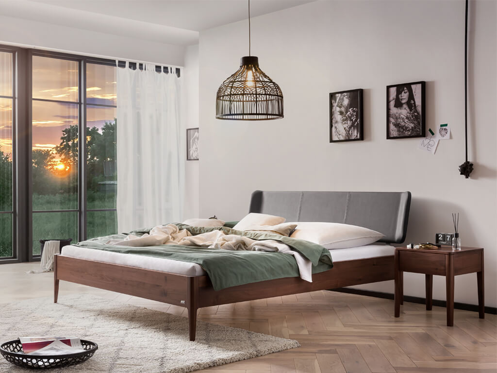 Hasena -  Lounge Bett Varese | Polsterkopfteil Boga | Holzfuß Pile | versch. Größen & Farben konfigurierbar Gerendertes Bild des Bettes Varese in einer dunklen Nussbaumausführung in einem Schlafzimmer, das ein großes Fenster und einen Nachttisch zeigt.