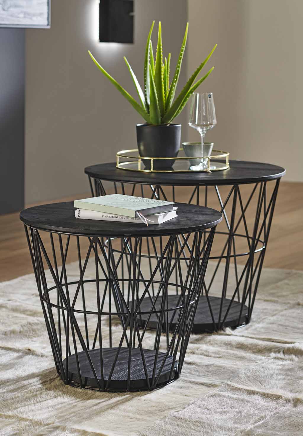 Zwei schwarze Couchtische mit Metallgestell, darauf eine Pflanze, ein Glas und Bücher, auf einem hellen Teppich platziert