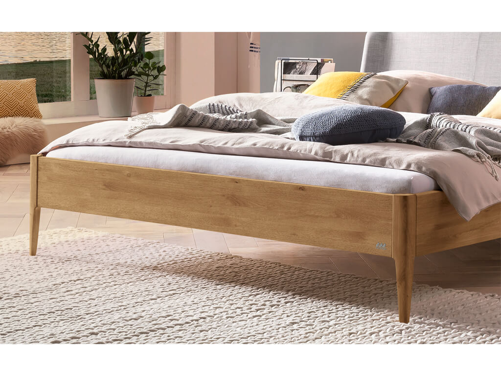 Eine Nahaufnahme des unteren Teils eines Bettes Pienza in Wildeiche, natur geölt. Das Bett ist auf schlanken, konischen Füßen platziert. Im Hintergrund sind ein Teppich und eine sonnendurchflutete Zimmerecke mit Pflanzen und einem Fenster zu sehen.