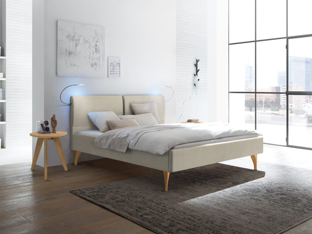 Ein beiges Polsterbett mit geteiltem Kopfteil und hellen Holzfüßen in einem modernen Raum mit einem Teppich.