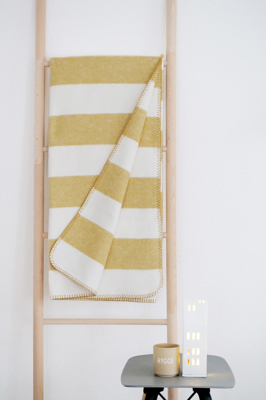 RIEMA. STINE – Wohndecke, Bio-Baumwolle - Streifen | 150 x 200 cm | gelb-weiß Gelb-weiß gestreifte Decke auf Holzleiter, begleitet von einem Hygge-Becher und einem kleinen, beleuchteten Dekorationsobjekt auf einem grauen Tisch.