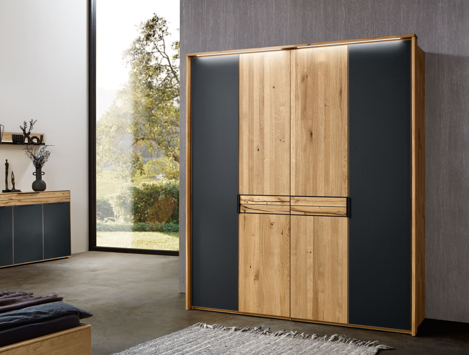 Wöstmann WSM 4300 - Kleiderschrank | Wildeiche massiv | optionale Mattglasfronten carbonfarbig | alle Breiten konfigurierbar Moderner Kleiderschrank aus Holz mit schwarzem Akzent in einem stilvollen Raum