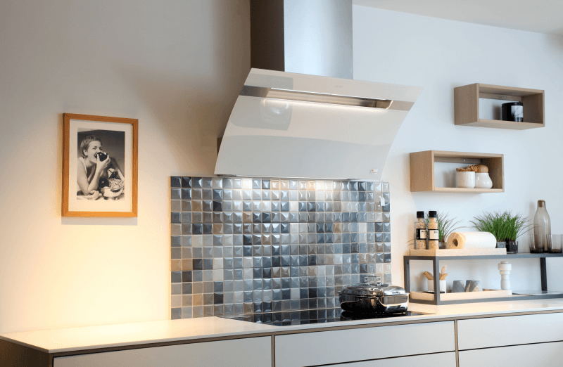 berbel - Kopffreihaube Glassline BKH 90 GL-2 Eine moderne Küche zeigt eine weiße schräge Dunstabzugshaube über einem schwarzen Induktionskochfeld und einem gefliesten Spritzschutz. Die Küche hat weiße Schränke, offene Holzregale und ein gerahmtes Bild an der Wand.