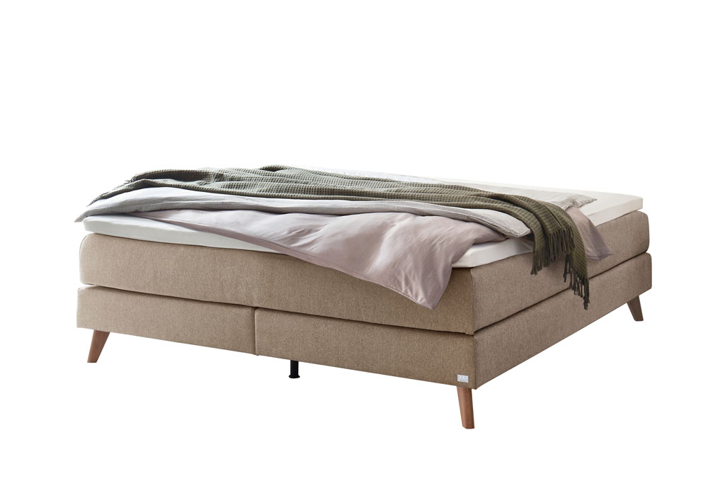 beiges Boxspringbett mit braunen Holzfüßen, stilvoll dekoriert mit Decken und Kissen vor weißem Hintergrund