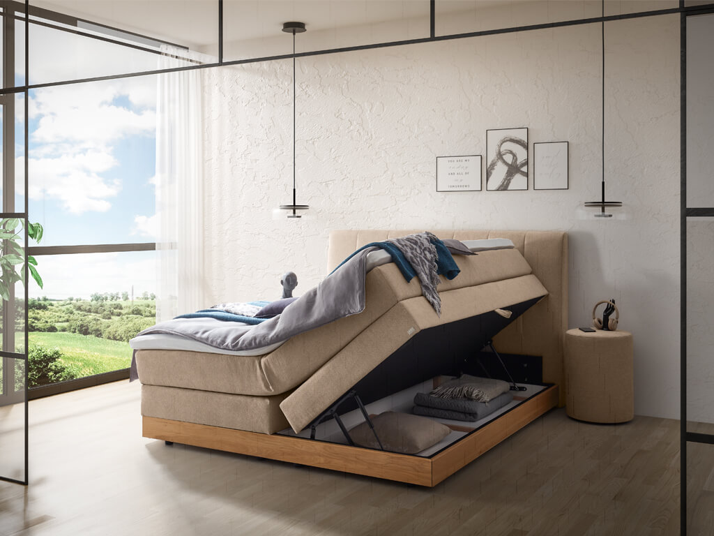 Ein beigefarbenes Boxspringbett in einem Schlafzimmer, dessen Matratze angehoben ist, um den Stauraum im Bettkasten zu zeigen.