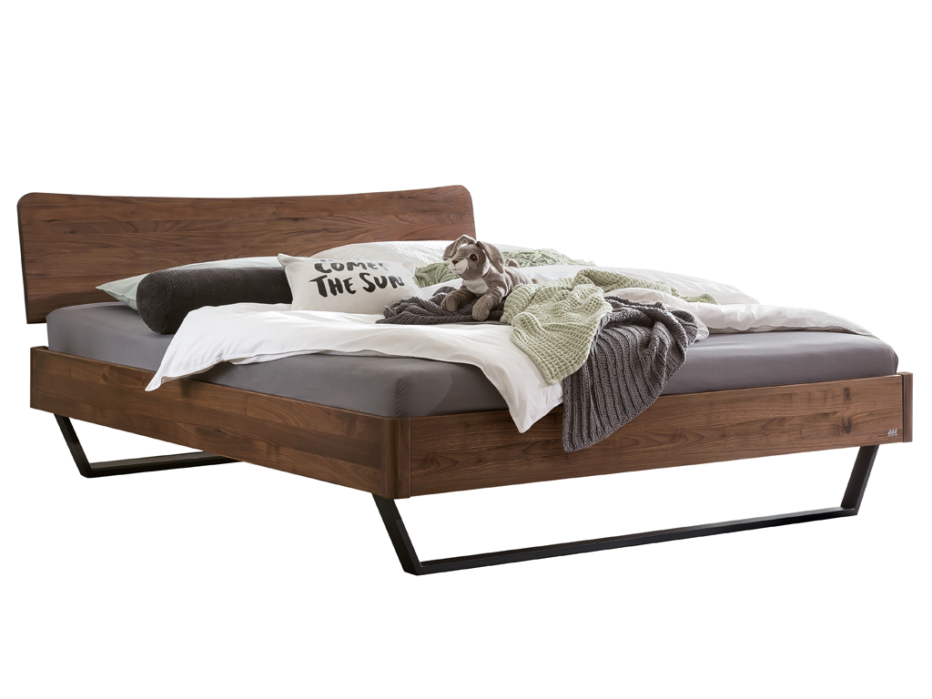 Ein Hasena Bettmodell Tresali aus Nussbaum mit einem geschwungenen Kopfteil und schwarzen Metallfüßen, auf dem eine Tagesdecke und Kissen liegen.