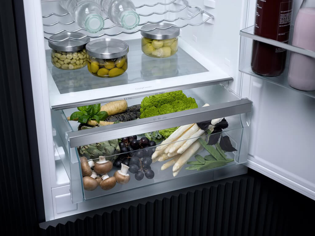 Das Bild zeigt die große Gemüseschublade eines Miele-Kühlschranks, gefüllt mit frischem Gemüse wie Spargel, Pilzen, Trauben, Salat und Artischocken.