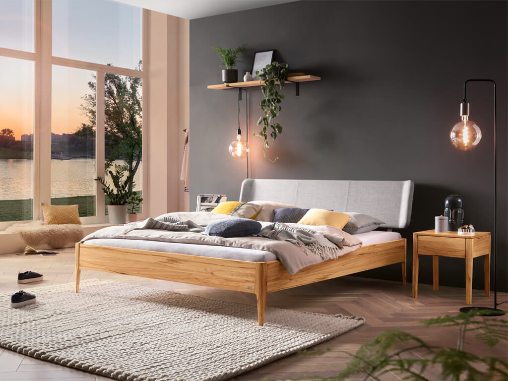 Ein gut beleuchteter Raum mit großen Fenstern und einem Hasena-Bett "Pienza Styly" aus Wildbuche. Das Bett ist mit einem gepolsterten, hellgrauen Kopfteil ausgestattet. Die Szene vermittelt eine warme und natürliche Atmosphäre.