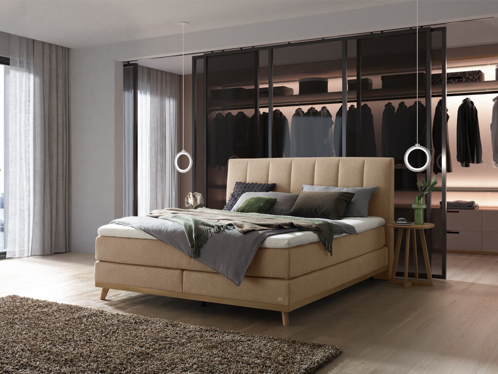 Ein Hasena Boxspringbett Pronto Stefania Roy mit sandfarbenem Stoffbezug, das in einem Schlafzimmer mit einem modernen begehbaren Kleiderschrank steht.
