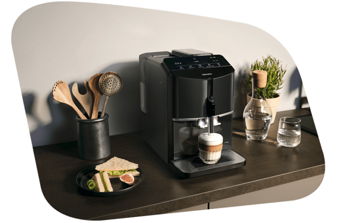 Ein schwarzer Siemens EQ Kaffeevollautomat brüht einen Latte Macchiato oder Cappuccino in eine Glastasse. Daneben steht ein Teller mit einem Sandwich und Küchenutensilien in einem Behältnis.