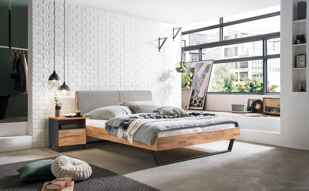 Hasena Trento - Bett Pietro | Kernbuche natur, geölt | Kopfteil Stoff grau Helles Schlafzimmer mit Holzbett, Dekokissen und modernen Leuchten vor einer Ziegelwand mit Fenster und Pflanzen.