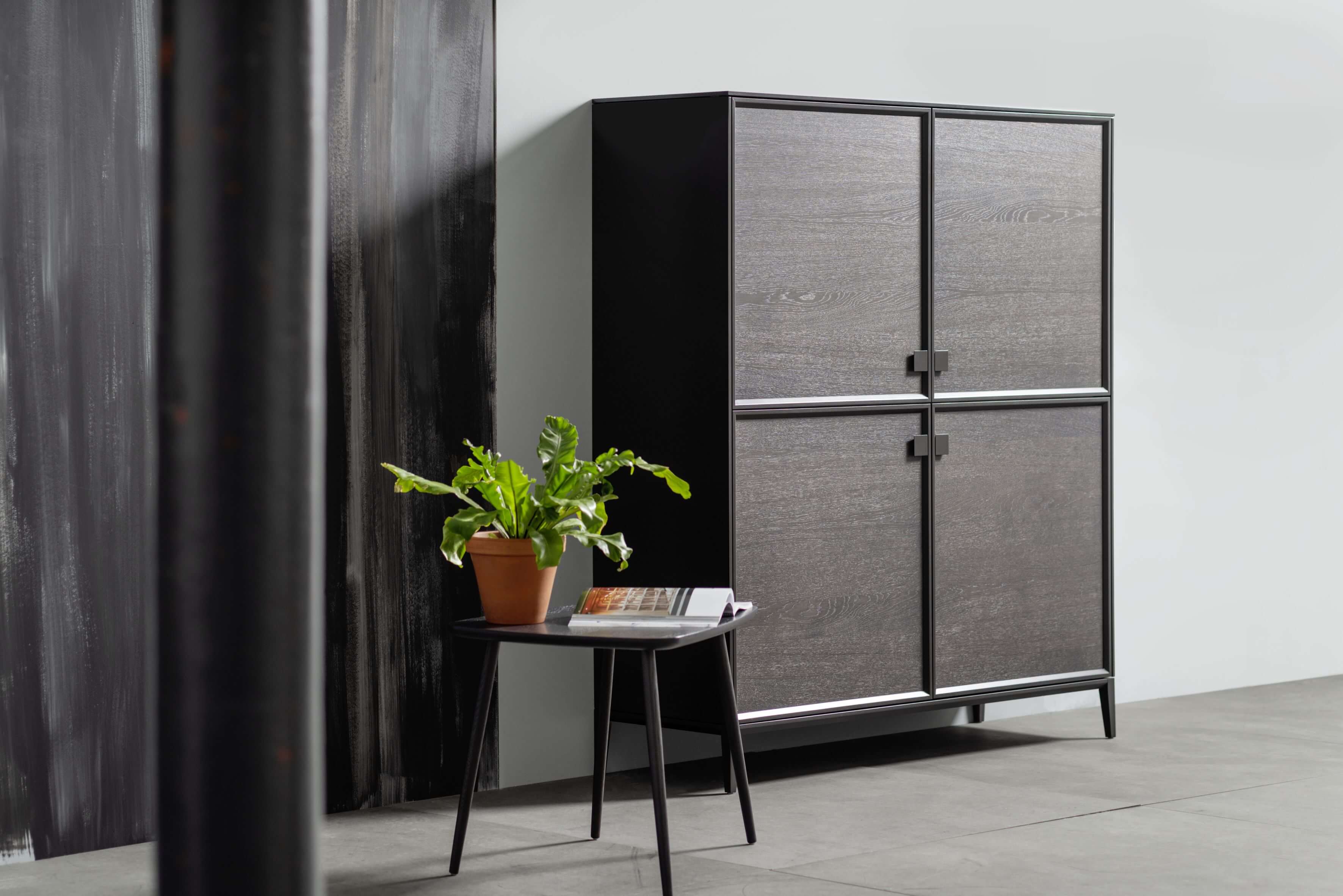 Dunkelbraunes Highboard mit minimalistischen Details und einem kleinen Beistelltisch mit einer Grünpflanze.