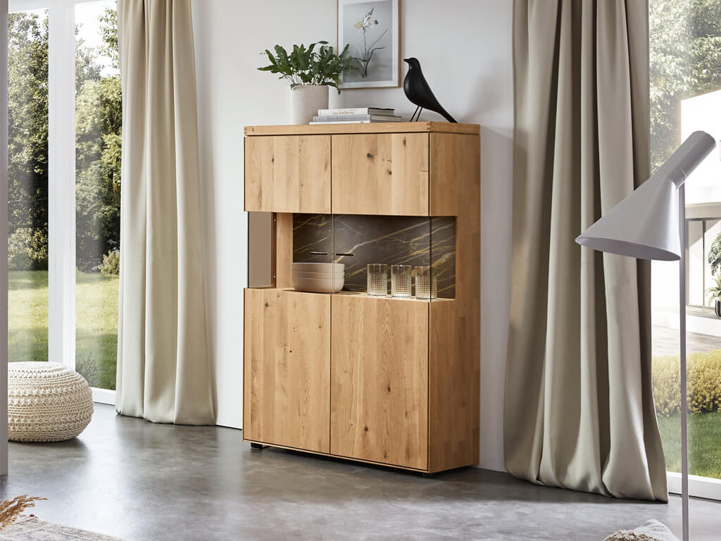 Highboard aus hellem Holz mit Holztüren und einem zentralen Glaseinsatz vor dunkler Rückwand. Das Schrankmöbel steht in einem lichtdurchfluteten Raum neben bodentiefen Fenstern mit beigen Vorhängen.
