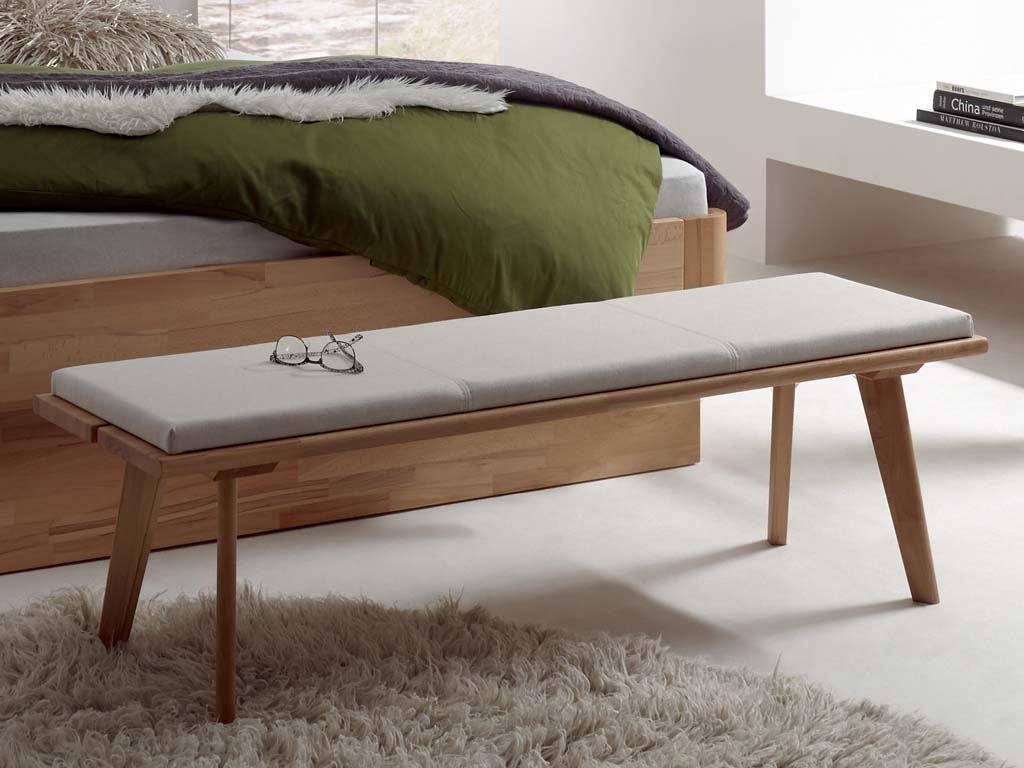moderne Holzbank in dunkelbraun mit einem Polsterkissen in grau wo eine Brille darauf liegt. auf einem grauen plüschteppich vor einem Bett