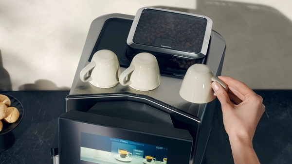 Hand platziert eine Tasse auf den Tassenwärmer einer Kaffeemaschine mit Bohnenbehälter und digitalem Display.