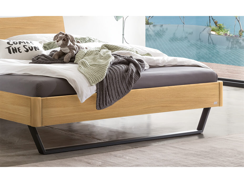 Nahaufnahme der Füße und des Bettrahmens vom Hasena Bett Tresali aus heller Eiche, das vor einem Fenster mit Pool-Blick steht.