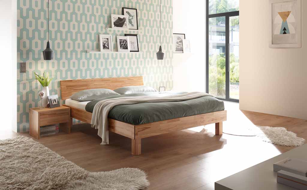 Hasena Trento - Bett Forti | Kernbuche natur, geölt Ein modernes Schlafzimmer mit einem Holzbett, grüner Bettwäsche und einer gemusterten Wand. Das Zimmer ist gut beleuchtet und stilvoll dekoriert.