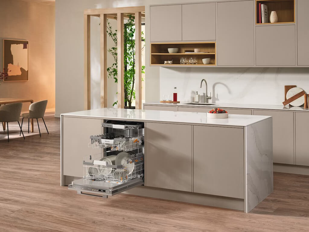 Eine moderne Küche mit einer geöffneten Miele Geschirrspülmaschine, die in die Kücheninsel integriert ist. Die Geschirrkörbe sind herausgezogen und mit Geschirr beladen.