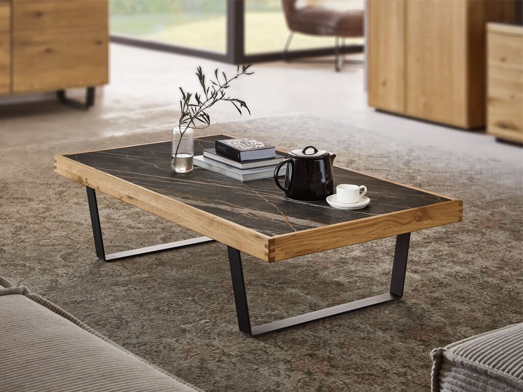 Rechteckiger Couchtisch aus Echtholz mit einer dunklen Platte in Steinoptik und schwarzen Metallkufen. Auf der Tischfläche stehen eine schwarze Kanne, eine Kaffeetasse, Bücher und eine kleine Vase.
