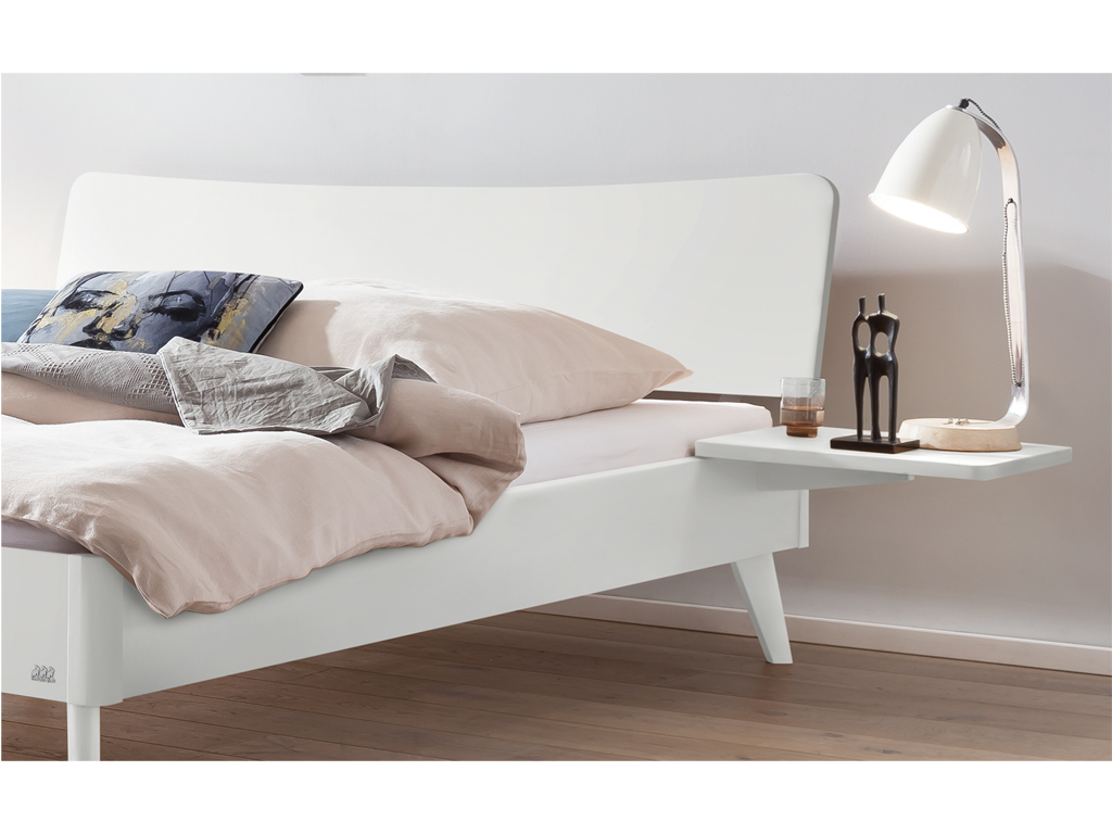 Ein weiß lackiertes Hasena Bett Caluso. Das Bett hat ein geschwungenes Kopfteil und schräg ausgestellte Beine. An der Seite ist ein schwebender Nachttisch mit einer Lampe und einer kleinen Skulptur zu sehen.
