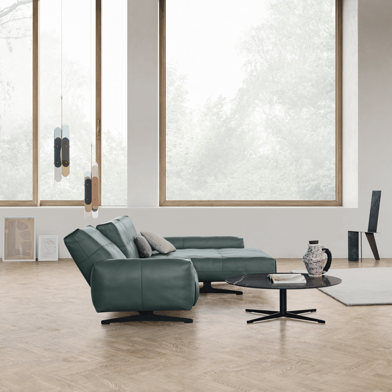 Das Bild zeigt ein modernes Wohnzimmer mit großen Fenstern, die viel natürliches Licht hereinlassen. Es gibt ein grünes L-förmiges Sofa von Rolf Benz 50 mit Kissen, einen runden schwarzen Couchtisch in der Mitte und einen grauen Teppich darunter. 