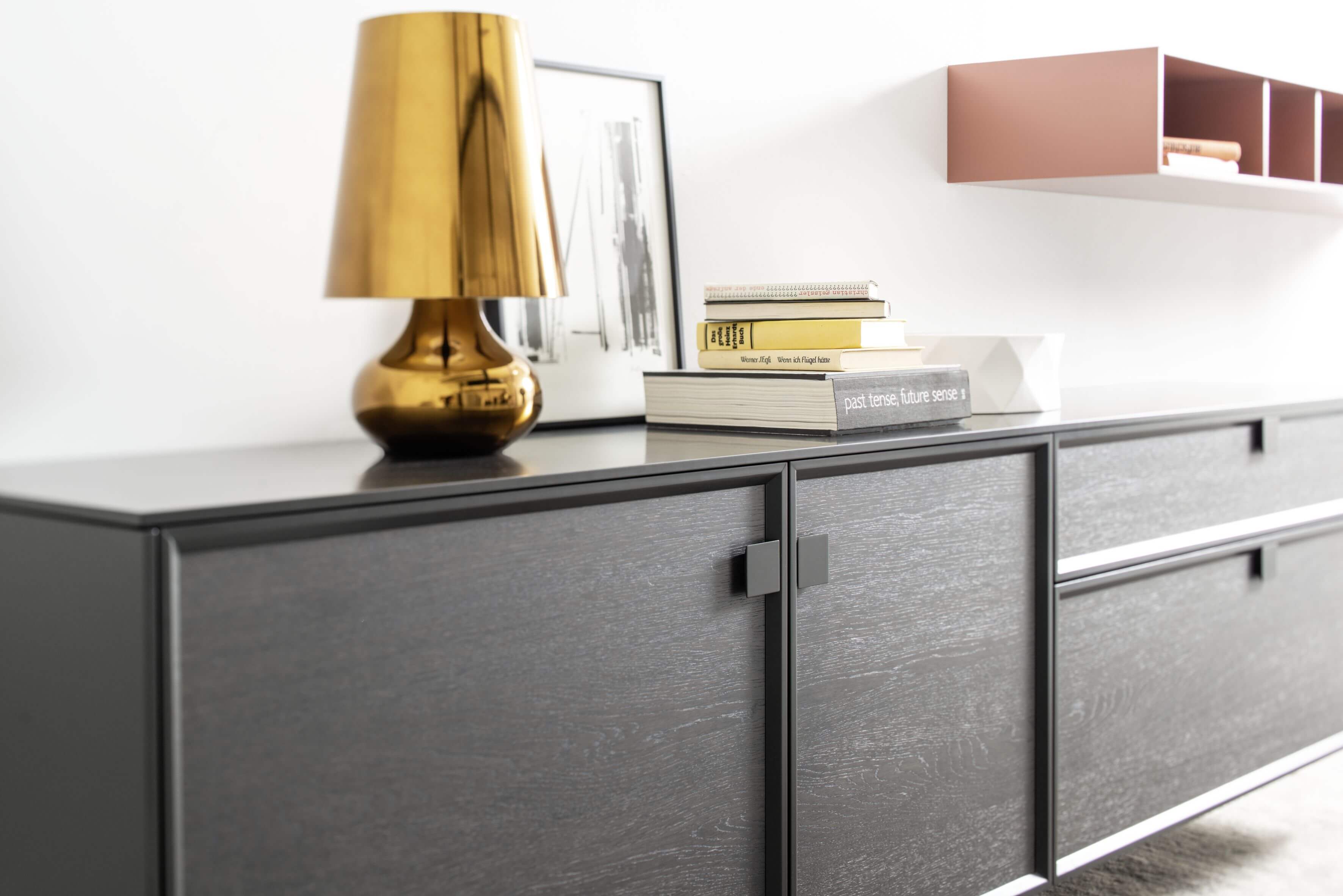 Das Bild zeigt ein modernes Interieur mit einem schwarzen Sideboard im Fokus. Auf dem Sideboard steht eine metallisch goldfarbene Tischlampe mit einem eleganten Design neben einem Stapel Hardcover-Bücher.