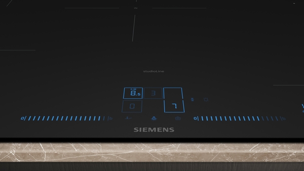 Nahaufnahme des unteren Bedienfelds eines Siemens StudioLine Induktionskochfelds. Man sieht die blauen Digitalanzeigen für die Leistungsstufen (z.B. "8.5" und "7") und die Slider-Bedienung auf der schwarzen Glaskeramikplatte.