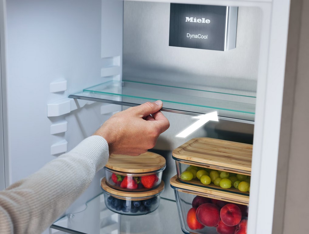 Eine Hand schiebt eine Glasablage in einem Miele Kühlschrank. Ein weißer Pfeil verdeutlicht die flexible Verstellmöglichkeit. Auf den unteren Ebenen stehen transparente Behälter mit frischen Beeren und Trauben.