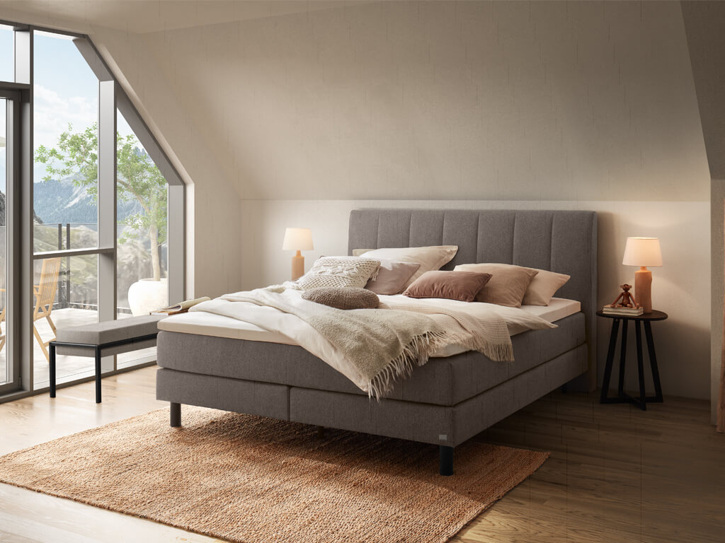 Ein Boxspringbett in einem Dachgeschosszimmer mit einem großen Fenster, das den Blick auf eine Berglandschaft freigibt.