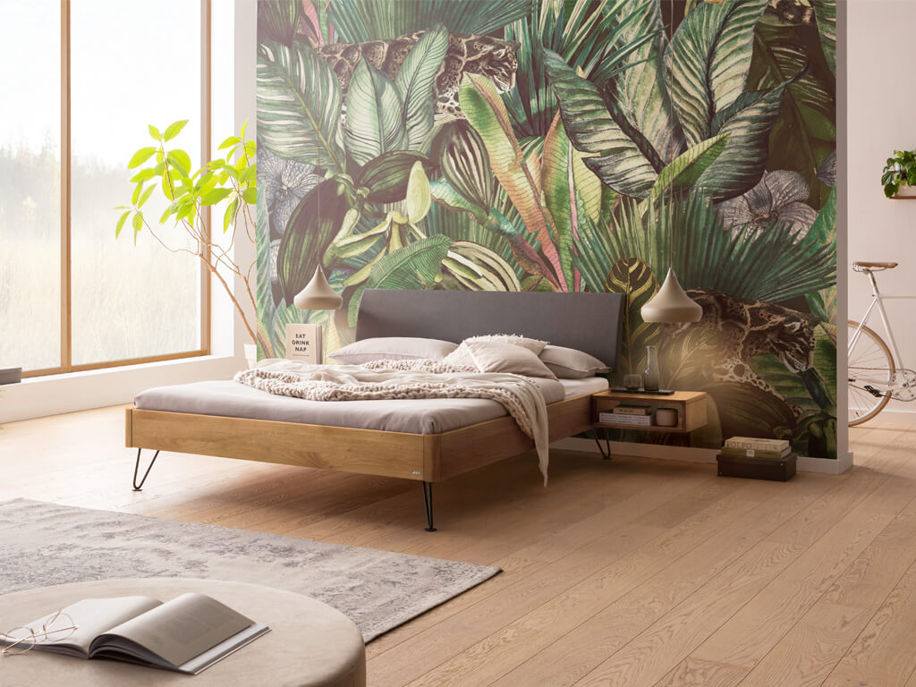 Hasena -  Lounge Bett Lugano | Polsterkopfteil Lecco | Metallfuß Locito | versch. Größen & Farben konfigurierbar Ein Schlafzimmer mit einem Bettrahmen aus naturbelassener geölter Wildeiche und einem anthrazitfarbenen Kopfteil. Die schwebenden Nachtkästchen sind ebenfalls aus Wildeiche. Der Raum ist mit einer Dschungel-Tapete und hellem Holzboden gestaltet.