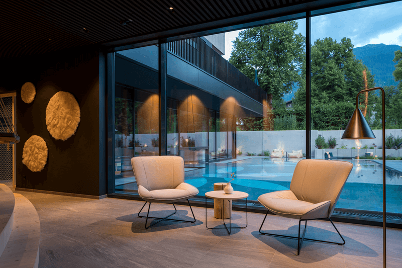 Elegante Lounge mit stilvollen Sesseln und Blick durch große Fenster auf den beleuchteten Pool im Garten