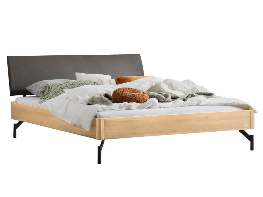 Eine Freisteller-Ansicht des Bettes "Broni" mit einem Bettrahmen aus heller Eiche. Es hat ein anthrazitfarbenes gepolstertes Kopfteil und steht auf schwarzen Metallfüßen. Das Bett ist mit weißen, grünen und orangefarbenen Kissen und Decken dekoriert.