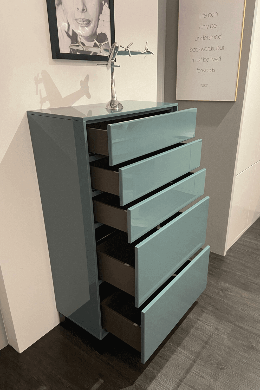 Eine moderne Kommode mit offenen Schubladen und glänzender Oberfläche, dekoriert mit einem Flugzeugmodell auf der Oberseite und gerahmten Kunstwerken an der Wand.