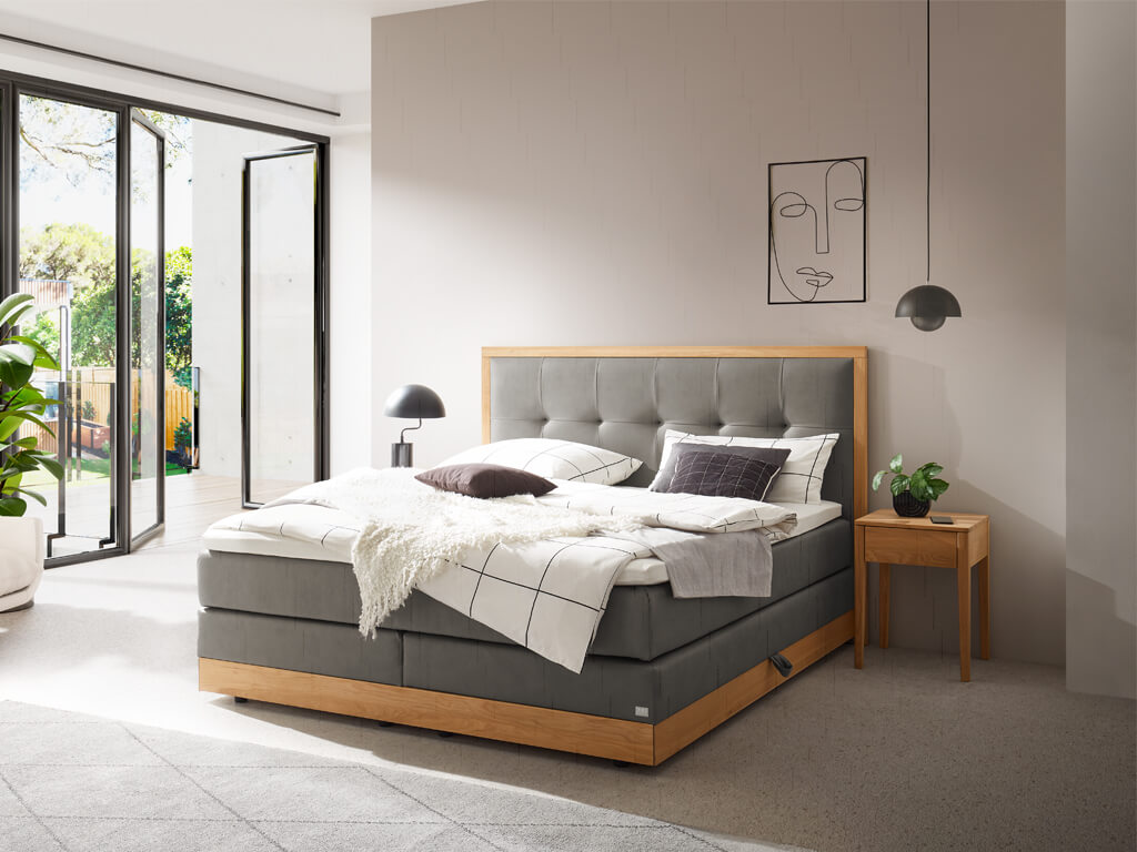 Modernes Boxspringbett "Hasena Pronto Alba" mit hellem Holzrahmen, grauem Stoffbezug und getuftetem Kopfteil. Weiße, graue und schwarze Bettwäsche, Kissen und eine weiße Decke.