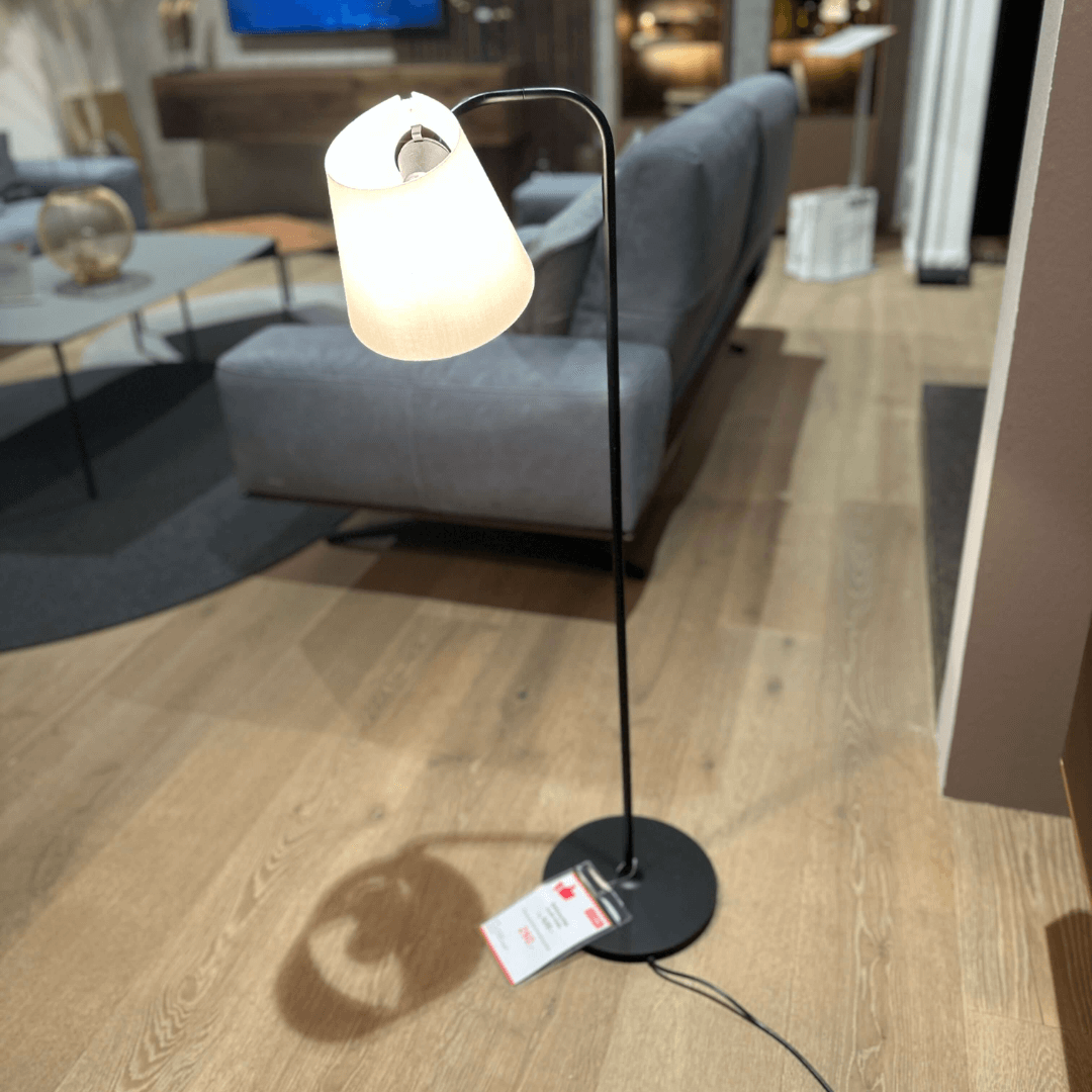 Eine moderne Stehlampe mit schlichtem, schwarzem Gestell und weißem Lampenschirm steht in einem Wohnzimmer mit Holzfußboden und grauem Sofa.