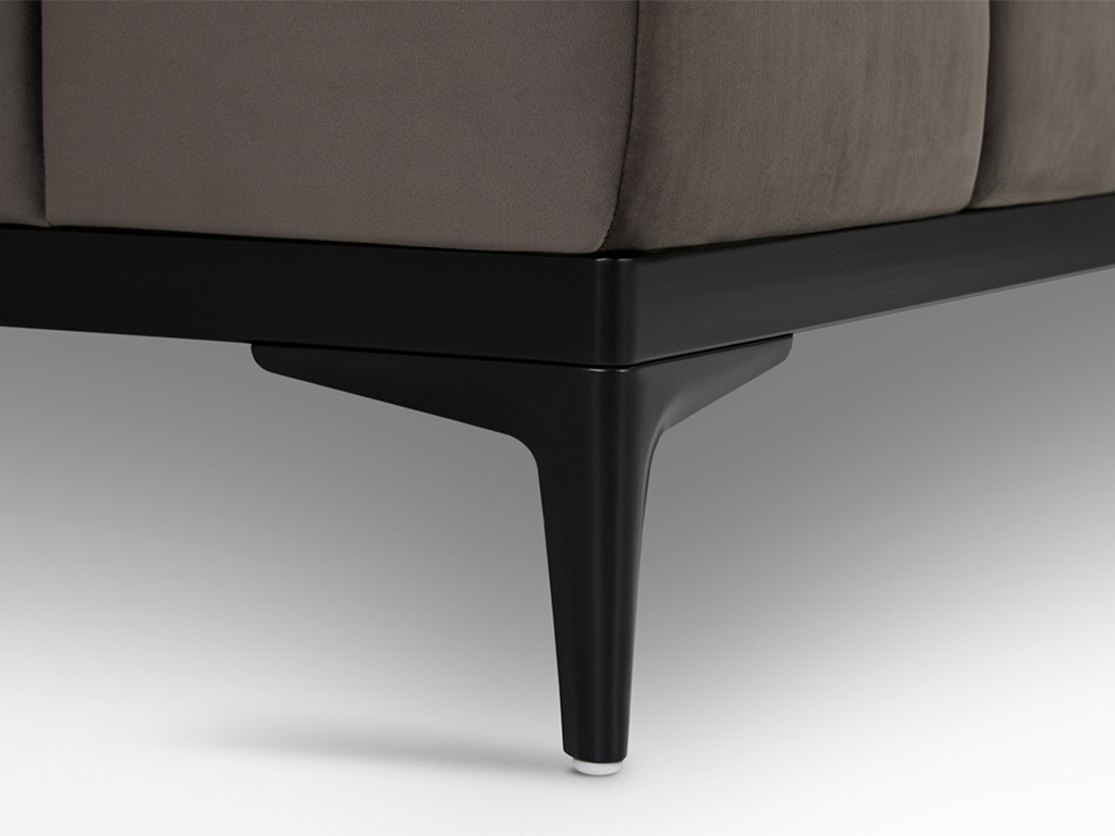 Nahaufnahme eines schwarzen, spitz zulaufenden Metallfußes eines Sofas. Der Fuß ist am Rahmen eines dunkelgrauen Sofamoduls befestigt und steht auf einem weißen Hintergrund.