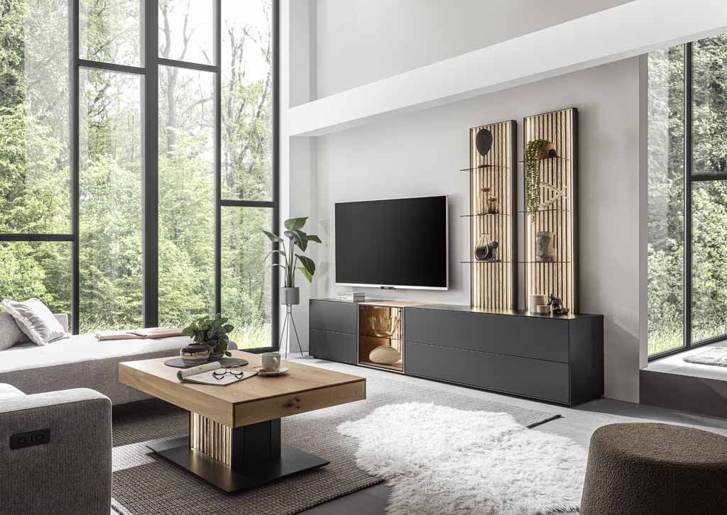 hartmann Selino - Wohnwand, 5-teilig | Type V28 | Birke/Kerneiche massiv, Lack anthrazit | B: 288,0 cm Ein helles Wohnzimmer mit großen Fenstern, einem modernen Fernseher und eleganten Möbeln, darunter ein Holztisch und schwarze Schränke.