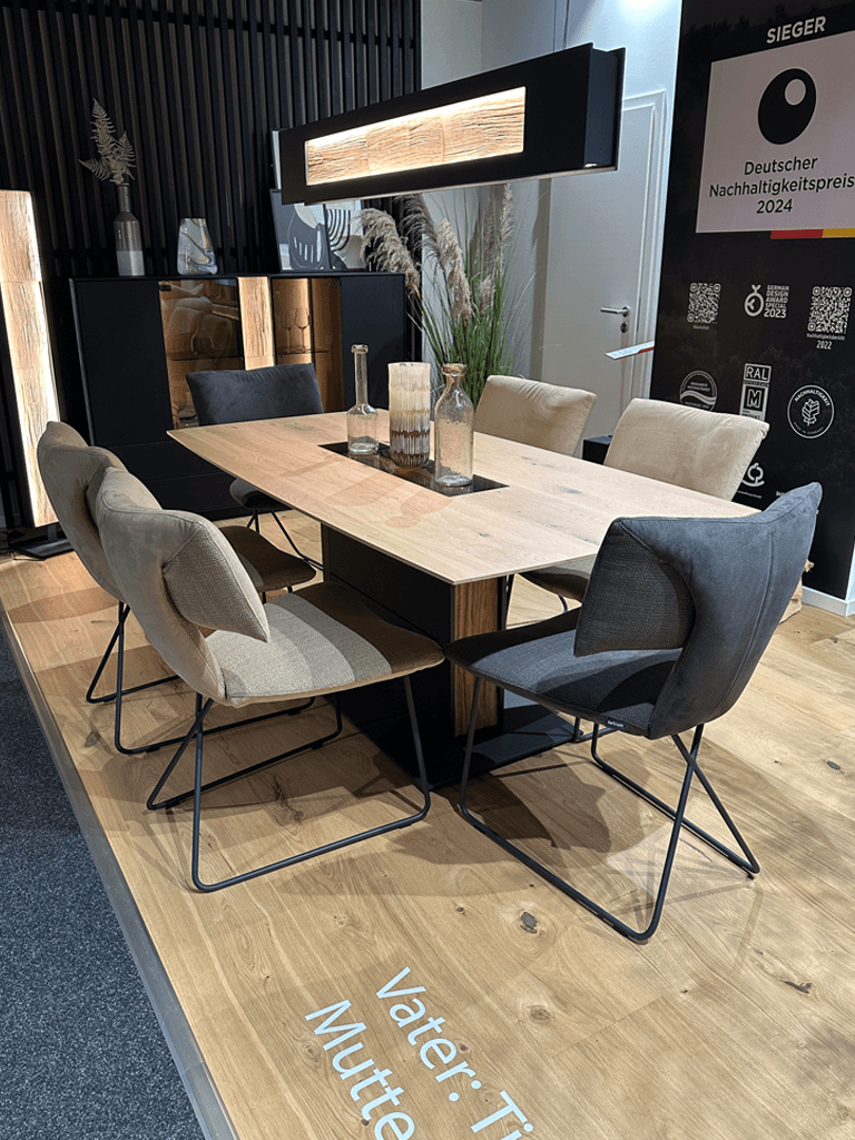 Hartmann KILVA | Essgruppe Moderner Essbereich mit einem eleganten Holz-Esstisch, umgeben von stilvollen Polsterstühlen. Eine atmosphärische Beleuchtung hängt über dem Tisch. Zimmerpflanzen und dekorative Elemente ergänzen das Design. Auszeichnung für Nachhaltigkeit im Hintergrund.