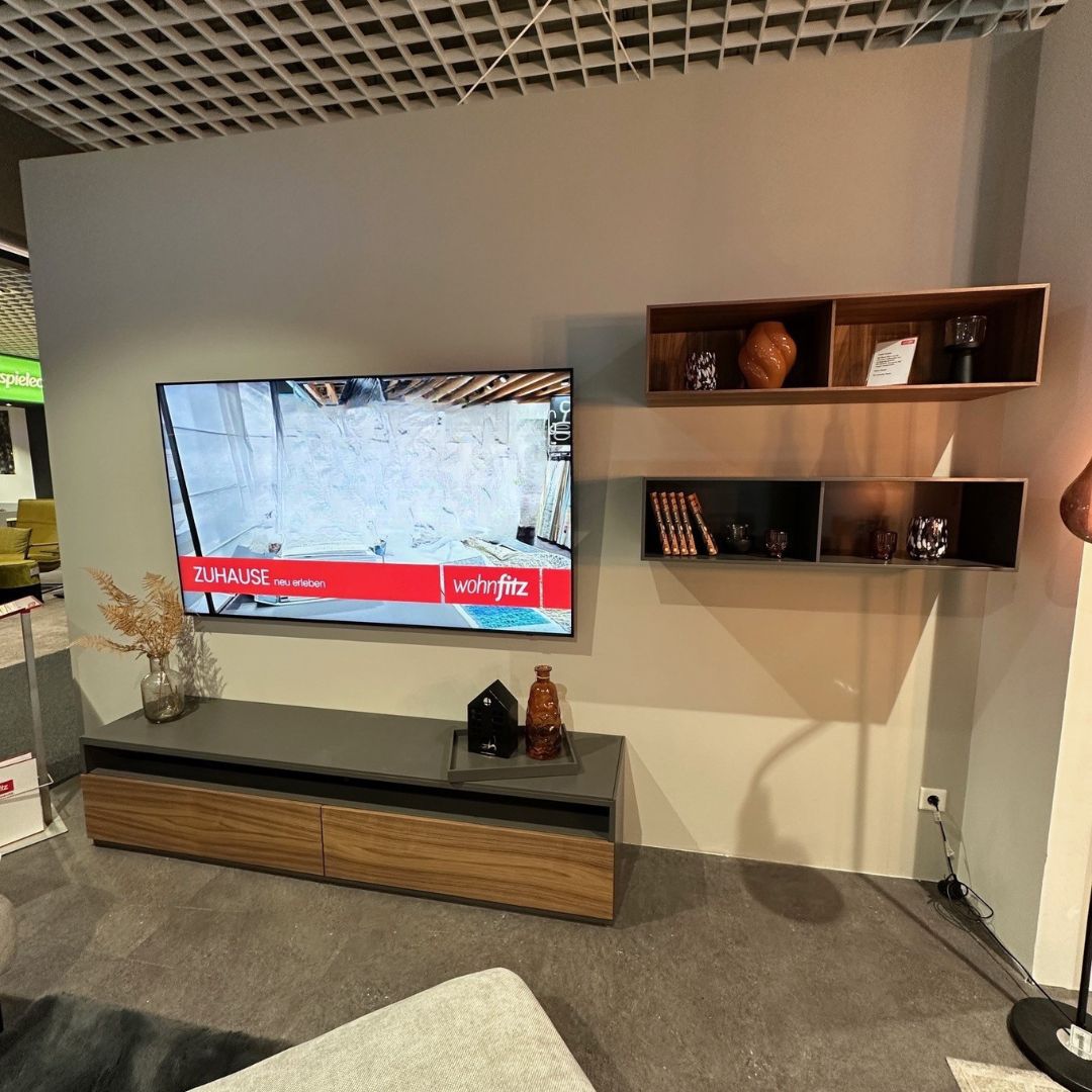 Rolf Benz STRETTO 9200 | Wohnkombination Ein modernes Wohnzimmer mit einem großen Flachbildfernseher an der Wand, stilvollem Holzmobiliar und dekorativen Regalen mit Vasen und Büchern.