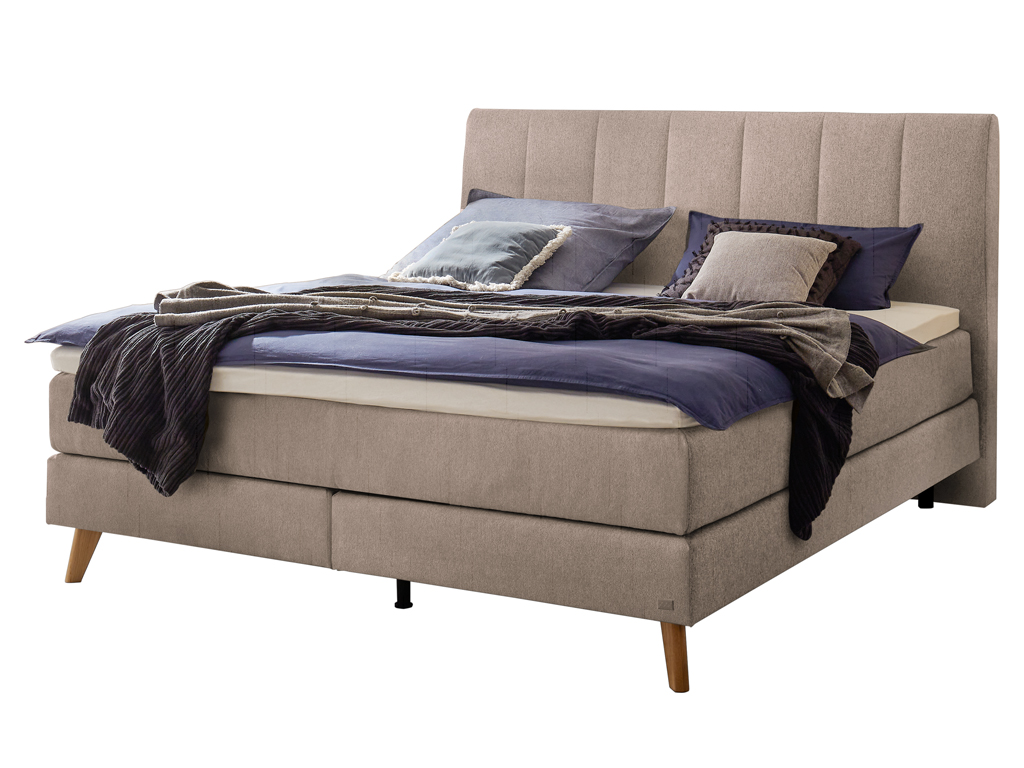 Ein Boxspringbett mit beigem Kopfteil, Holzfüßen und passender Matratze und Bettwäsche vor einem weißen Hintergrund.
