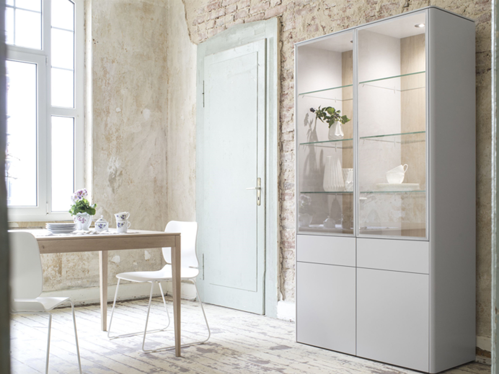 Sudbrock. Goya - Vitrine | Lack steingrau, Eiche milk Furnier | 4 Türen, 2 Schubladen | B: 125,2 cm Ein modernes Wohnzimmer mit einer Glasvitrine, gefüllt mit Geschirr und Dekorationen, neben einem kleinen Esstisch mit weißer Tischdecke und Blumenarrangement. Der Raum hat eine helle, minimalistische Einrichtung.