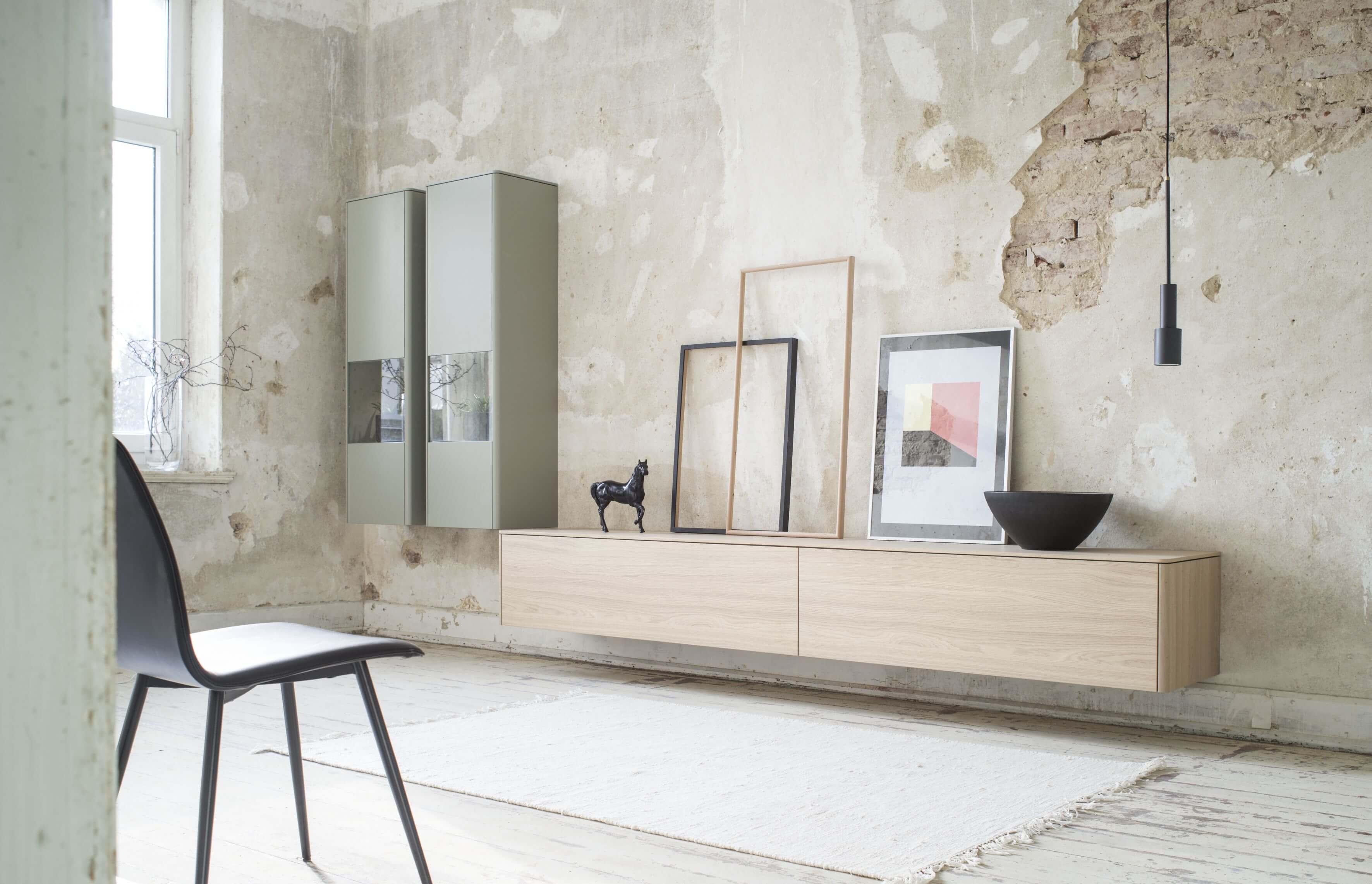 Sudbrock Goya - Wohnwand | Lowboard und 2 Hängevitrinen | Lack salbei, Eiche milk | B: 387,8 cm Ein modern eingerichtetes Wohnzimmer mit schwebendem Sideboard, Dekorstücken und einem schwarzen Stuhl vor einer unvollendeten Betonwand.