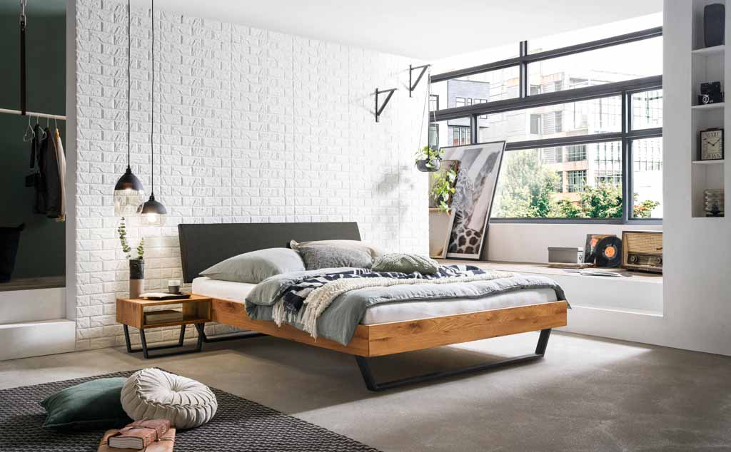 Hasena Trento - Bett Sariano | 140 x 200 cm | Wildeiche natur, geölt | Kopfteil Kunstleder grau Stilvolles Schlafzimmer mit einem Doppelbett aus Holz, Natur- und Weißtönen, dekorativen Elementen und großen Fenstern mit Blick nach draußen