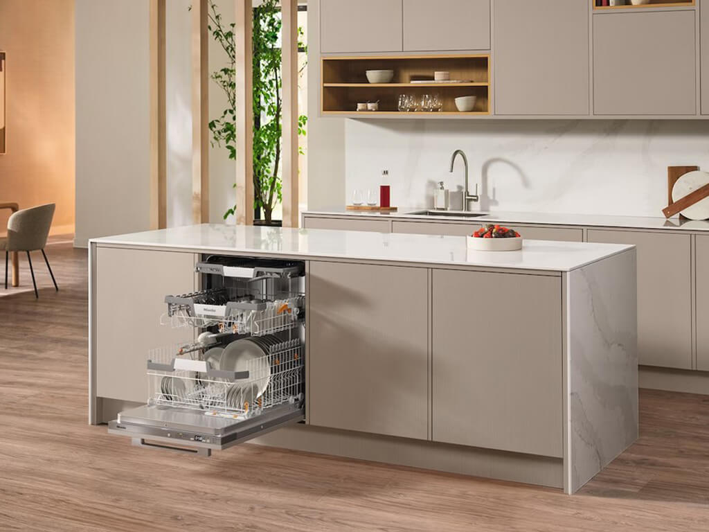 Eine moderne Küche mit einer geöffneten Miele Geschirrspülmaschine, die in die Kücheninsel integriert ist. Die Geschirrkörbe sind herausgezogen und mit Geschirr beladen.